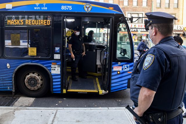 Two MTA officers distribute masks on a bus whose LED sign says "Masks required